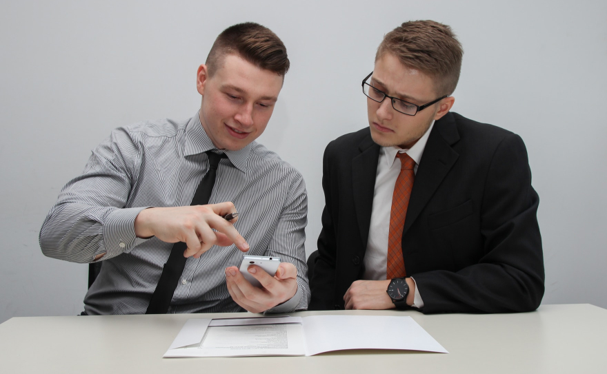 Two young businessmen in a meeting discussing a contract. One man is holding a smartphone and pointing to something on the screen, while the other man is listening attentively.