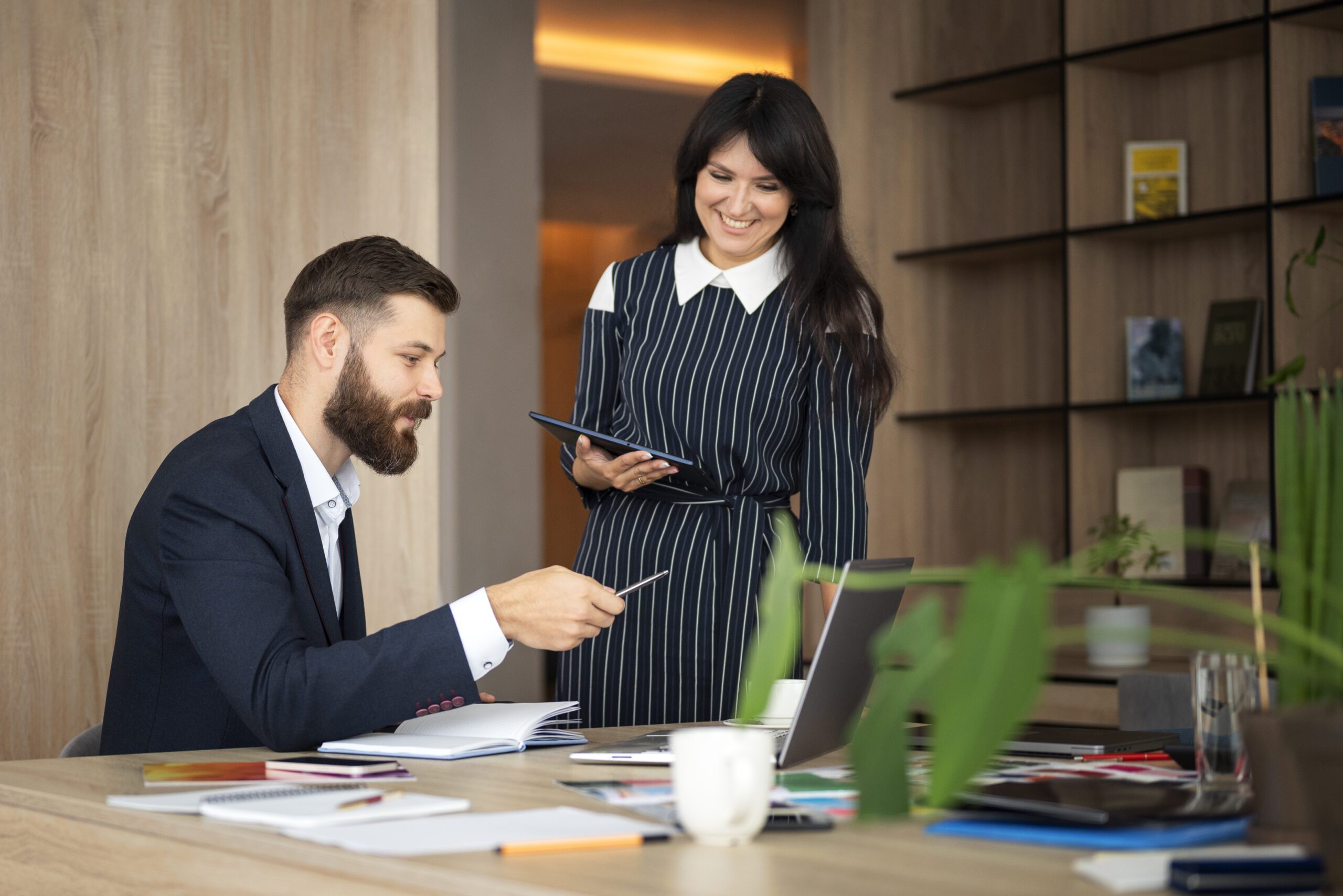 Man and woman collaborating at a desk; the man points at a notebook while the woman smiles, holding a tablet.